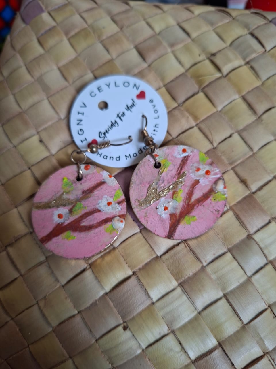 A pair of pink hand painted earrings showing blossom on a branch, on a woven mat